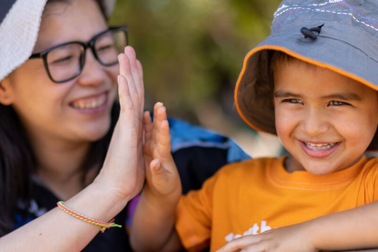An Early Childhood Teacher (ECT) at a preschool is giving a high-five to a young child, both wearing sun hats and smiling brightly